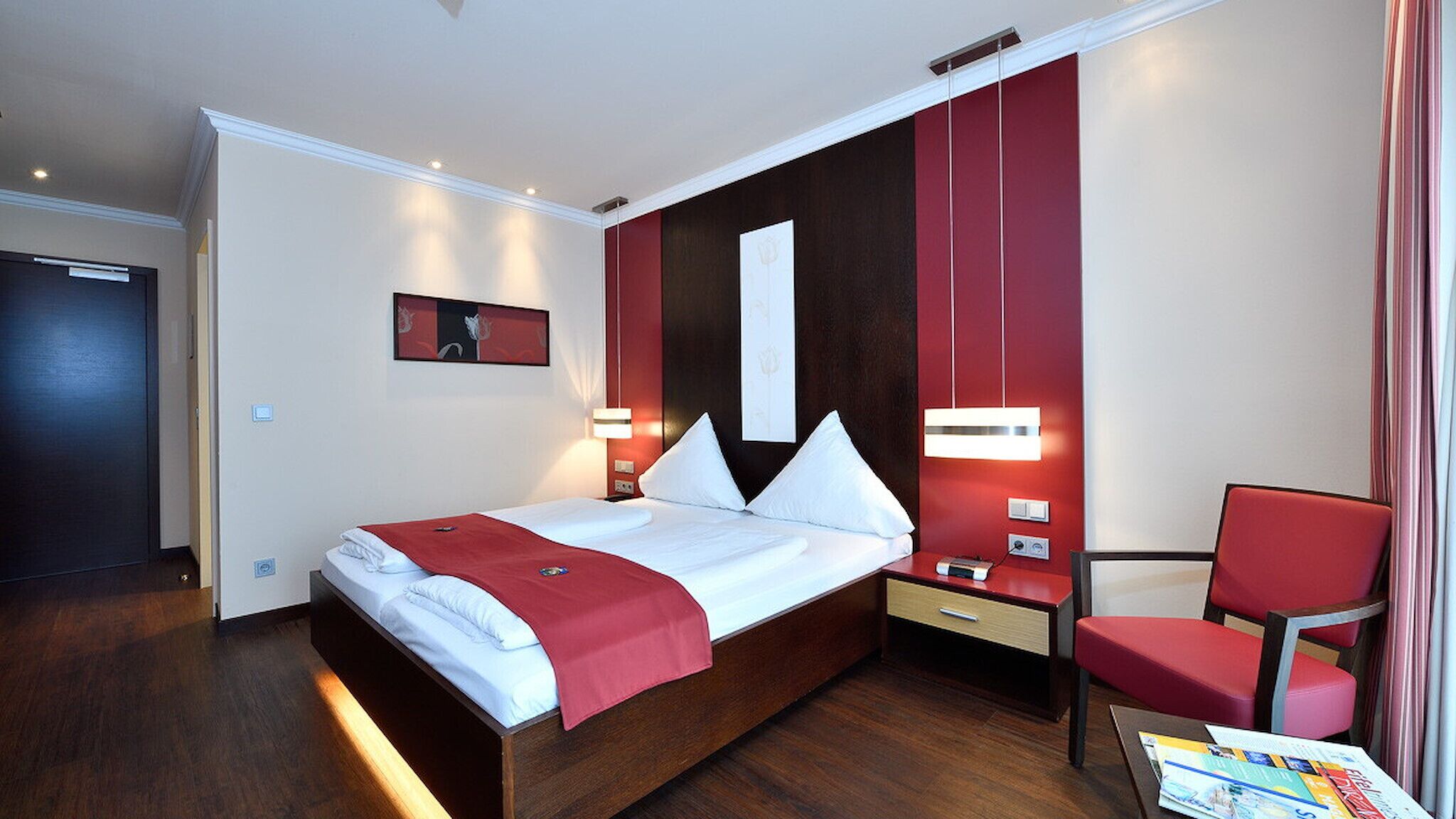 Una habitación de hotel moderna con una cama grande, una pared de acento roja, una silla roja y una mesita de noche con lámpara.