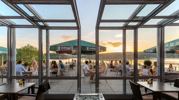 A restaurant with outdoor seating, large windows, and a view of a building with a green roof. - Dormio Hotel der Seehof (Heimbach)