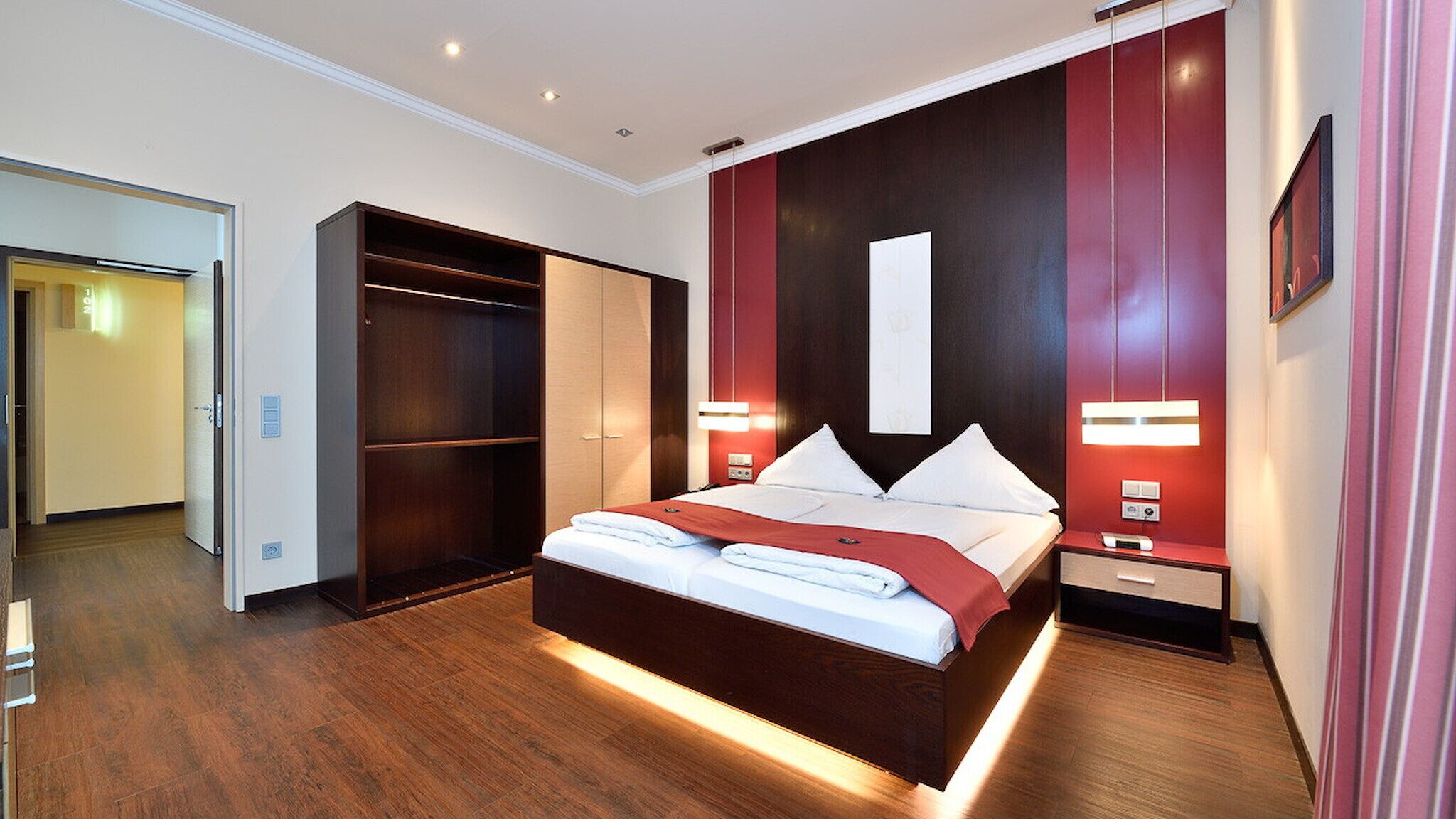 A modern hotel room with a large bed, built-in wardrobe, bedside tables, and a large mirror.