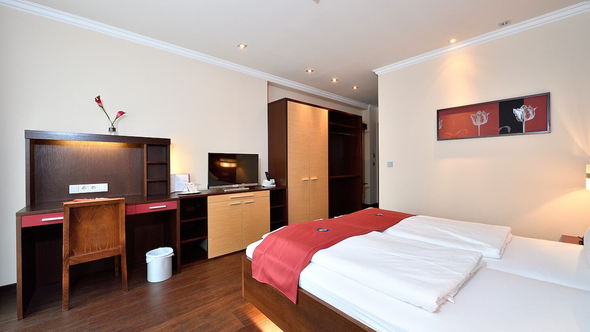 Ein Hotelzimmer mit Bett, Schreibtisch mit Computer, Stuhl, Kleiderschrank und einem Bild an der Wand.