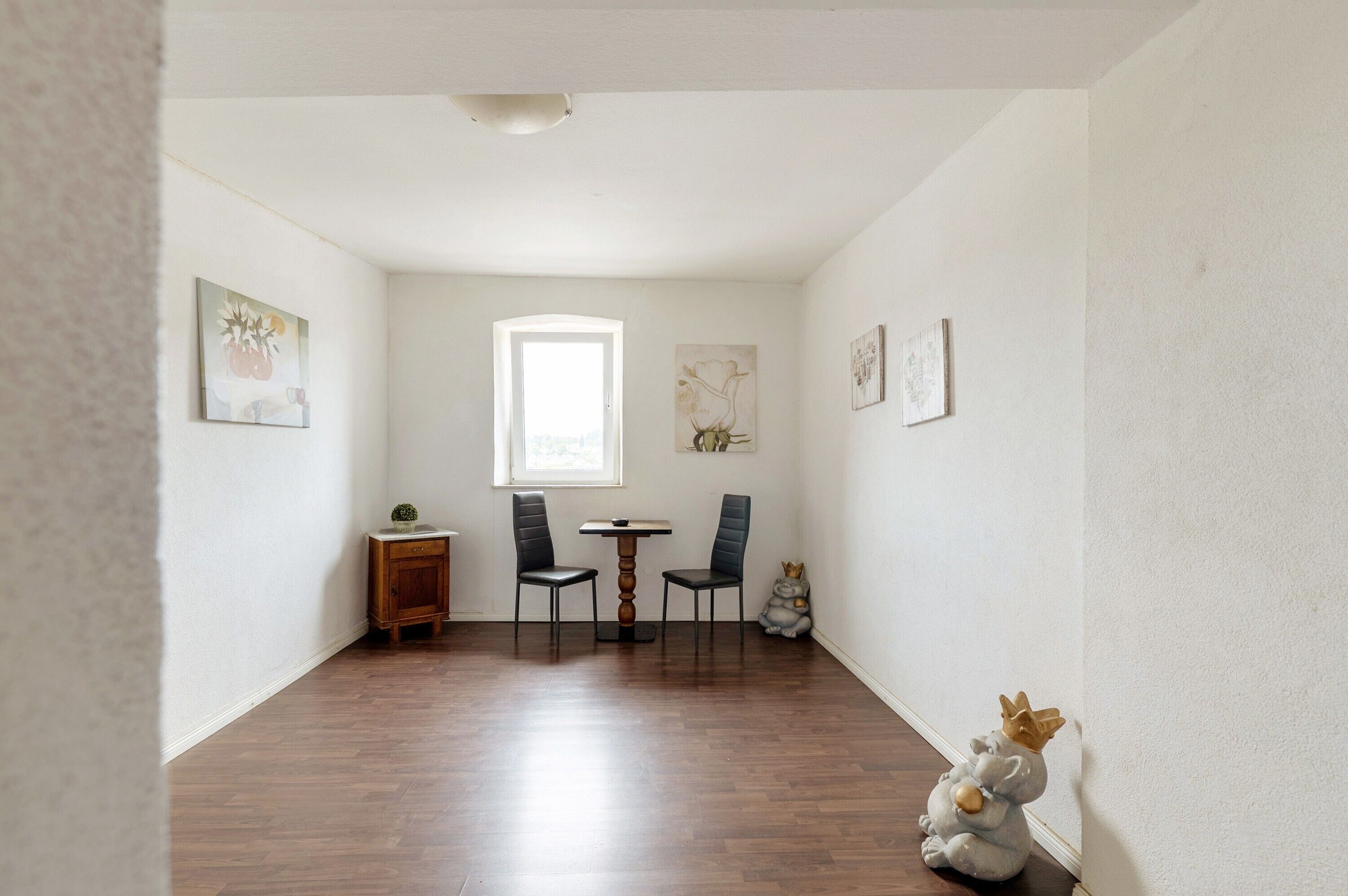 A room with a wooden floor, a small table with two chairs, a cabinet, and a window.
