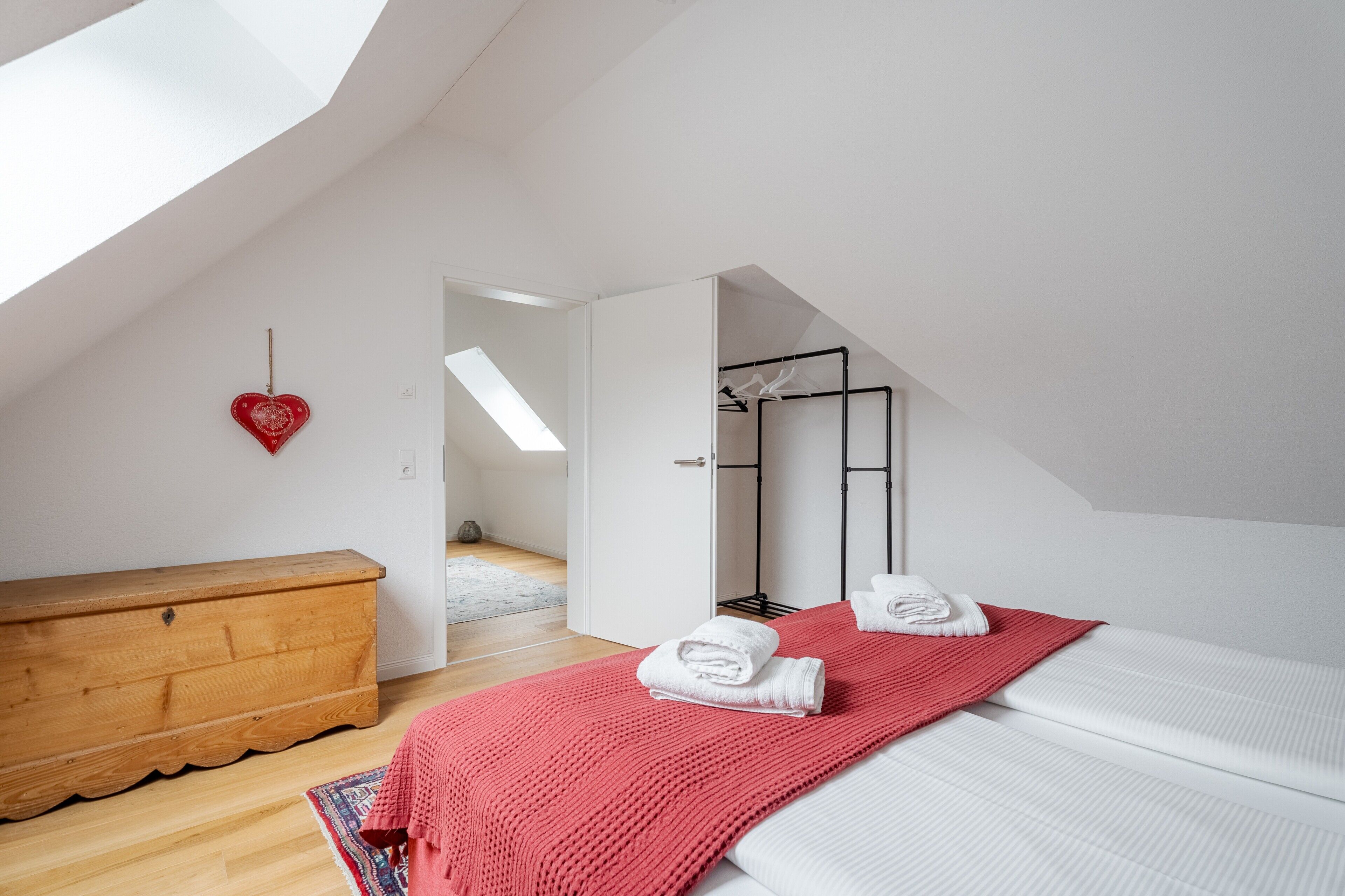 Une chambre avec un banc en bois, une couverture rouge, un lit blanc, une décoration en forme de cœur et une lucarne.