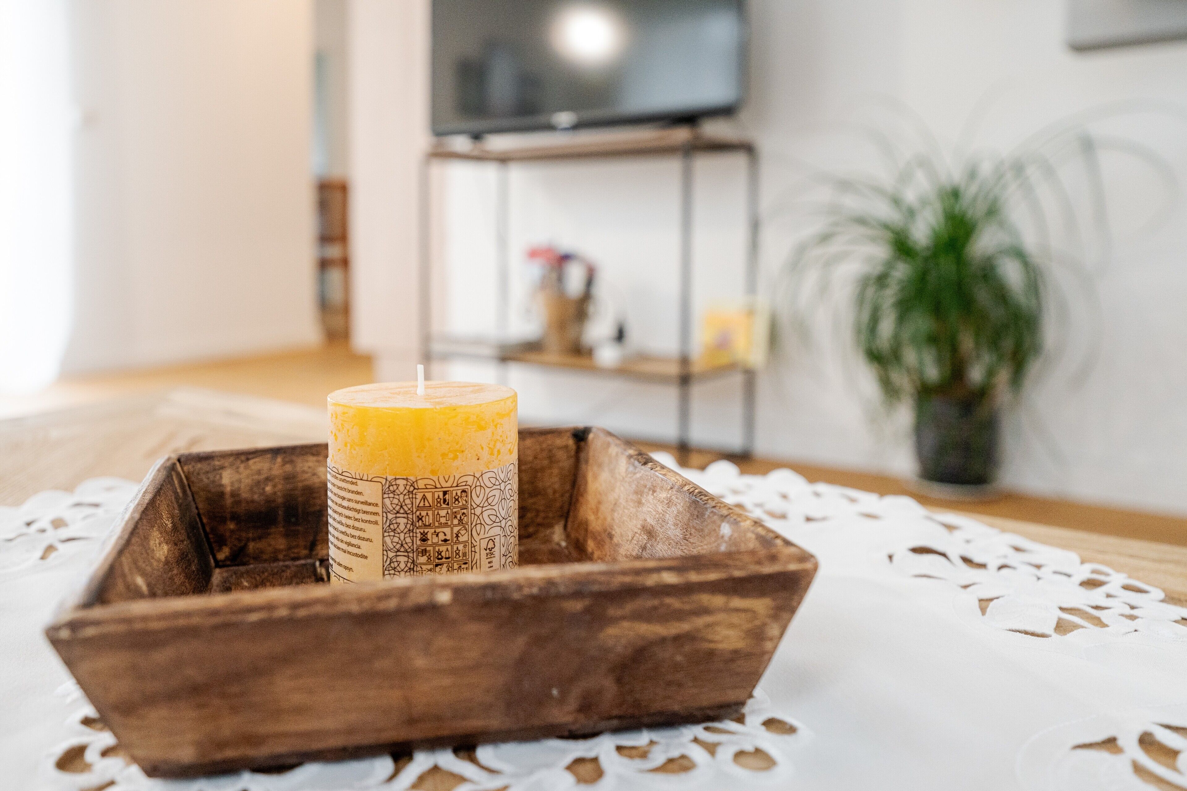 Une bougie dans un plateau en bois, posée sur une table recouverte d’un chemin de table, avec une télévision et une plante en arrière-plan.