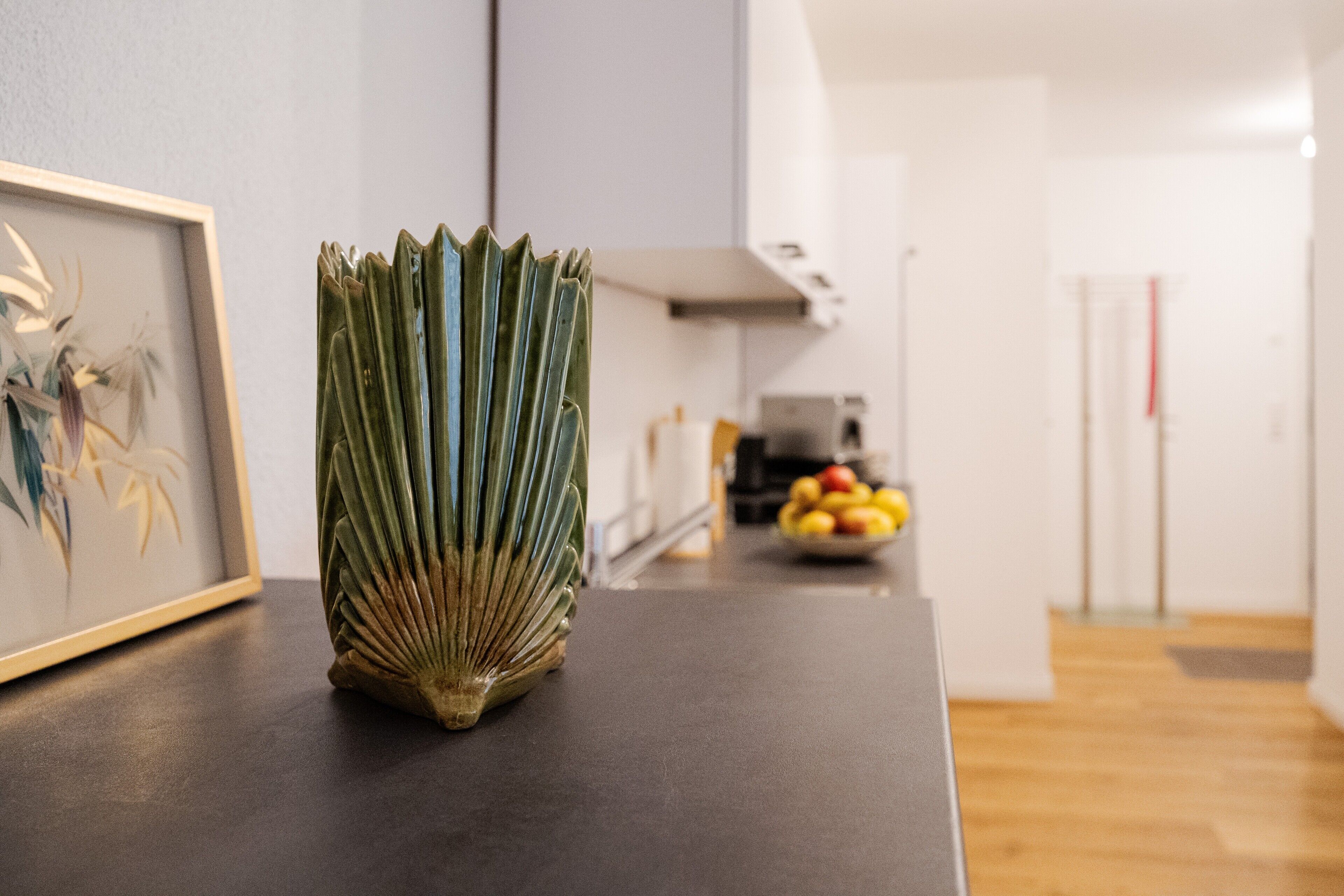 Eine Küche mit einem gerahmten Kunstwerk, einem grünen Strauß und einer Obstschale im Hintergrund.