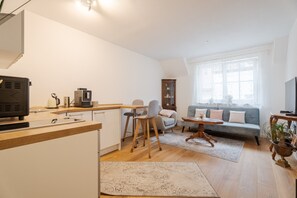 A modern kitchen with a built-in oven, a dining area with a wooden table and chairs, a gray sofa with a patterned rug, and a large window with sheer curtains.