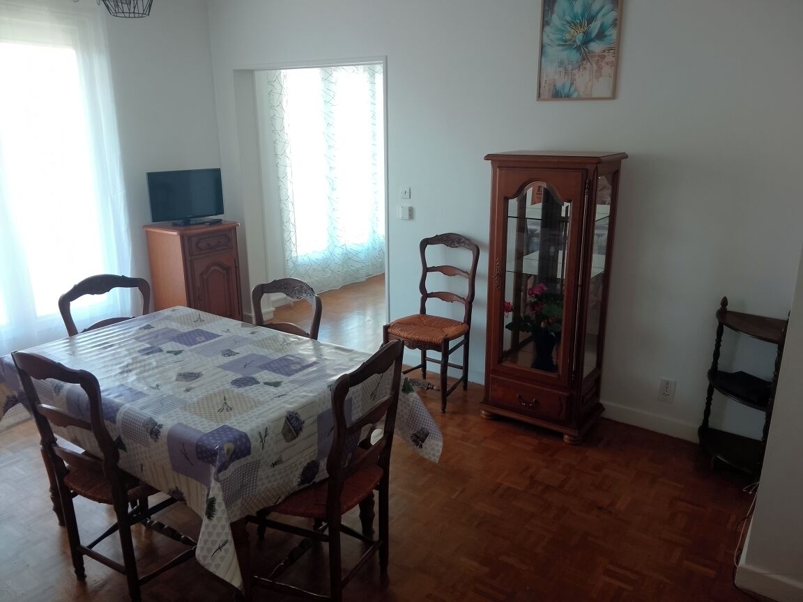 Ein Essbereich mit einem Tisch, der mit einer gemusterten Tischdecke bedeckt ist, umgeben von Holzstühlen. Im Hintergrund ist ein Holzschrank mit Glastüren zu sehen.