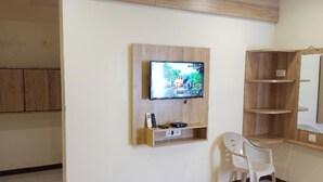 A room with a mounted TV, wooden shelves, a chair, and a wooden cabinet. - Hotel Radhika (Somnath)