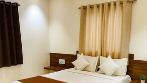 A neatly made bed with a wooden headboard, a bedside table with a lamp, and a window with curtains. - Hotel Radhika (Somnath)
