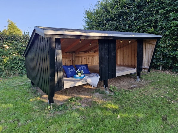 Eine hölzerne Konstruktion mit überdachter Veranda, einer Bank mit Kissen und einem kleinen Tisch mit Tablett.