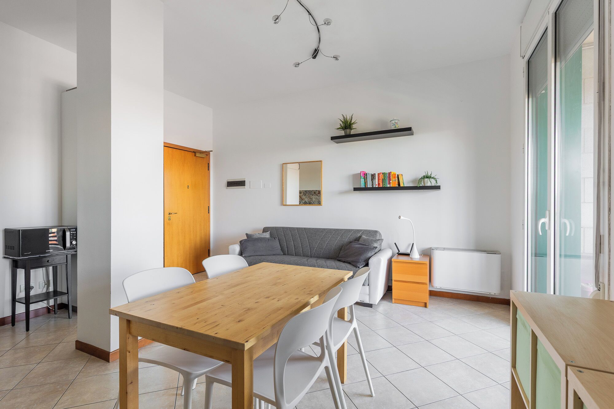 Una sala de estar moderna con una mesa de comedor de madera, sillas blancas, un sofá gris, una pequeña estantería y un radiador.
