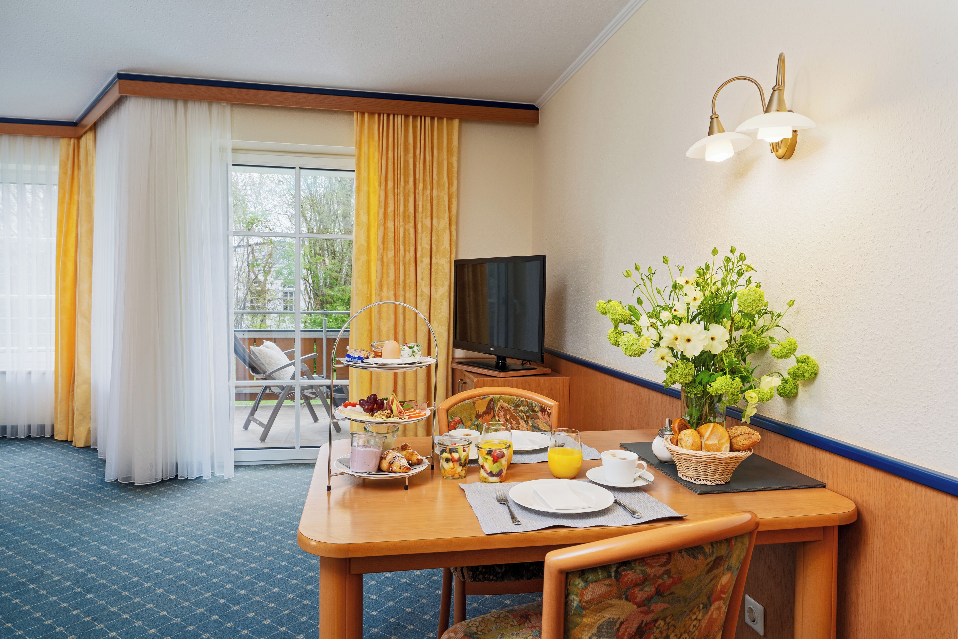 Ein Hotelzimmer mit Esstisch, gedecktem Frühstücksteller, Fernseher und einem Balkon mit gelben Vorhängen.