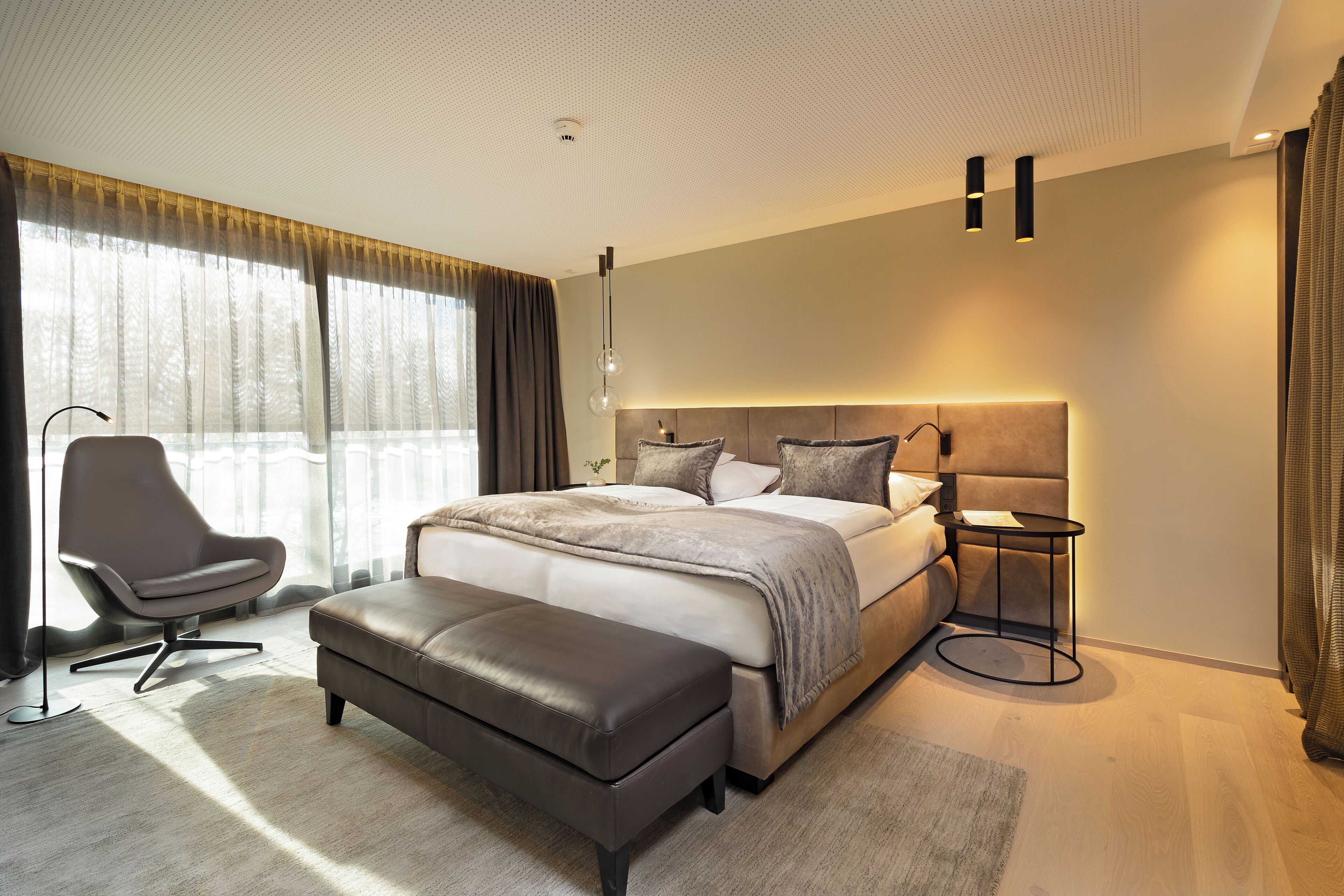 A modern bedroom with a large bed, a side table, a chair, and a large window with curtains.