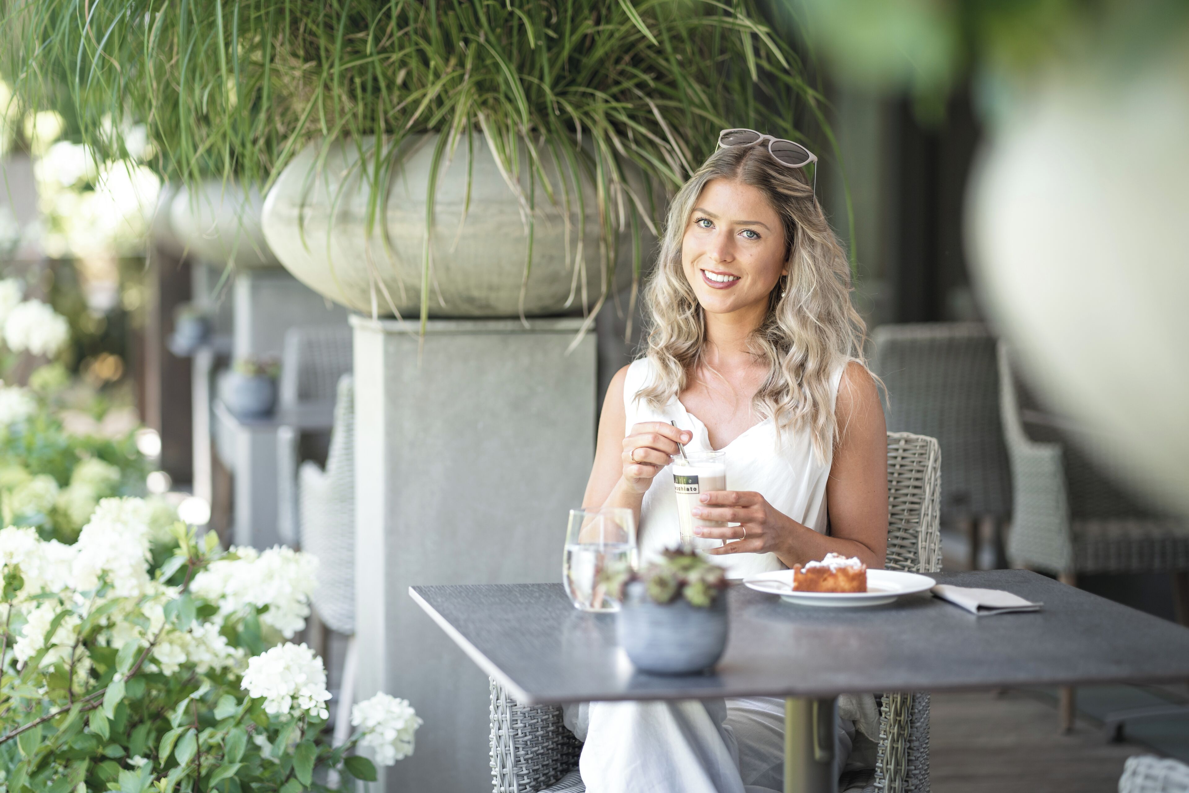 Una persona sentada en una mesa al aire libre, con una bebida y un plato de comida, rodeada de follaje y plantas.