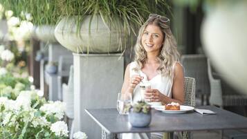 Una persona sentada en una mesa al aire libre, con una bebida y un plato de comida, rodeada de follaje y plantas.