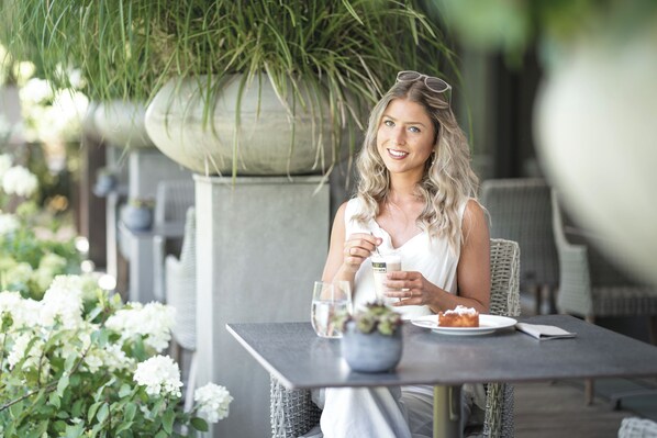 Une personne est assise à une table en plein air, avec une boisson et une assiette de nourriture devant elle, entourée de verdure et de plantes.