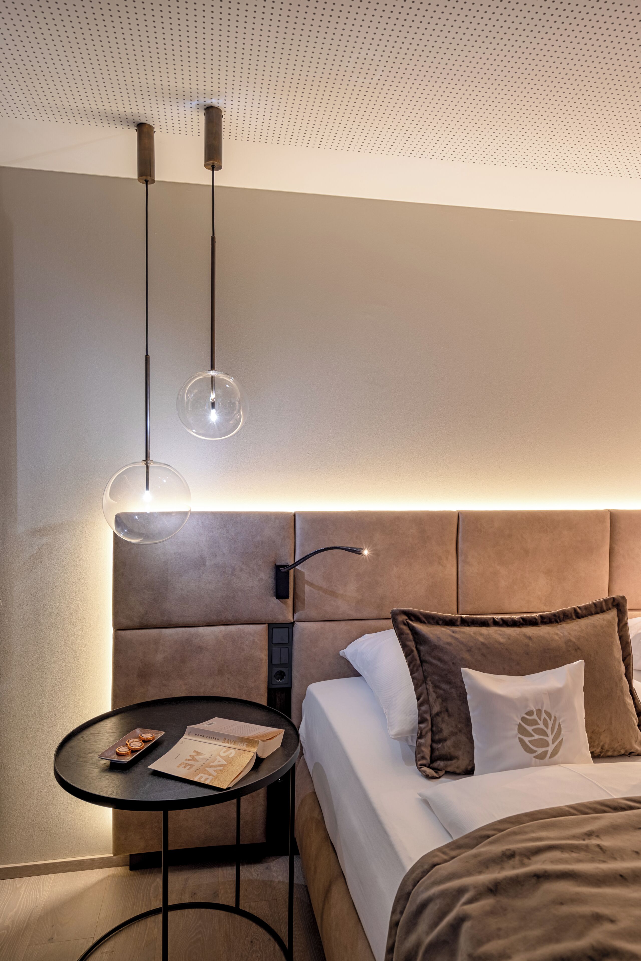 Una habitación de hotel moderna con una cama grande, una mesita redonda y dos lámparas colgantes.