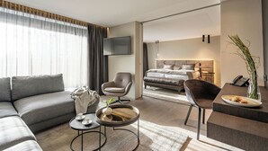 A modern hotel room with a sofa, armchair, coffee table, and a dining area with a vase and plate. - Hotel Riedenburg (Bad Fuessing)