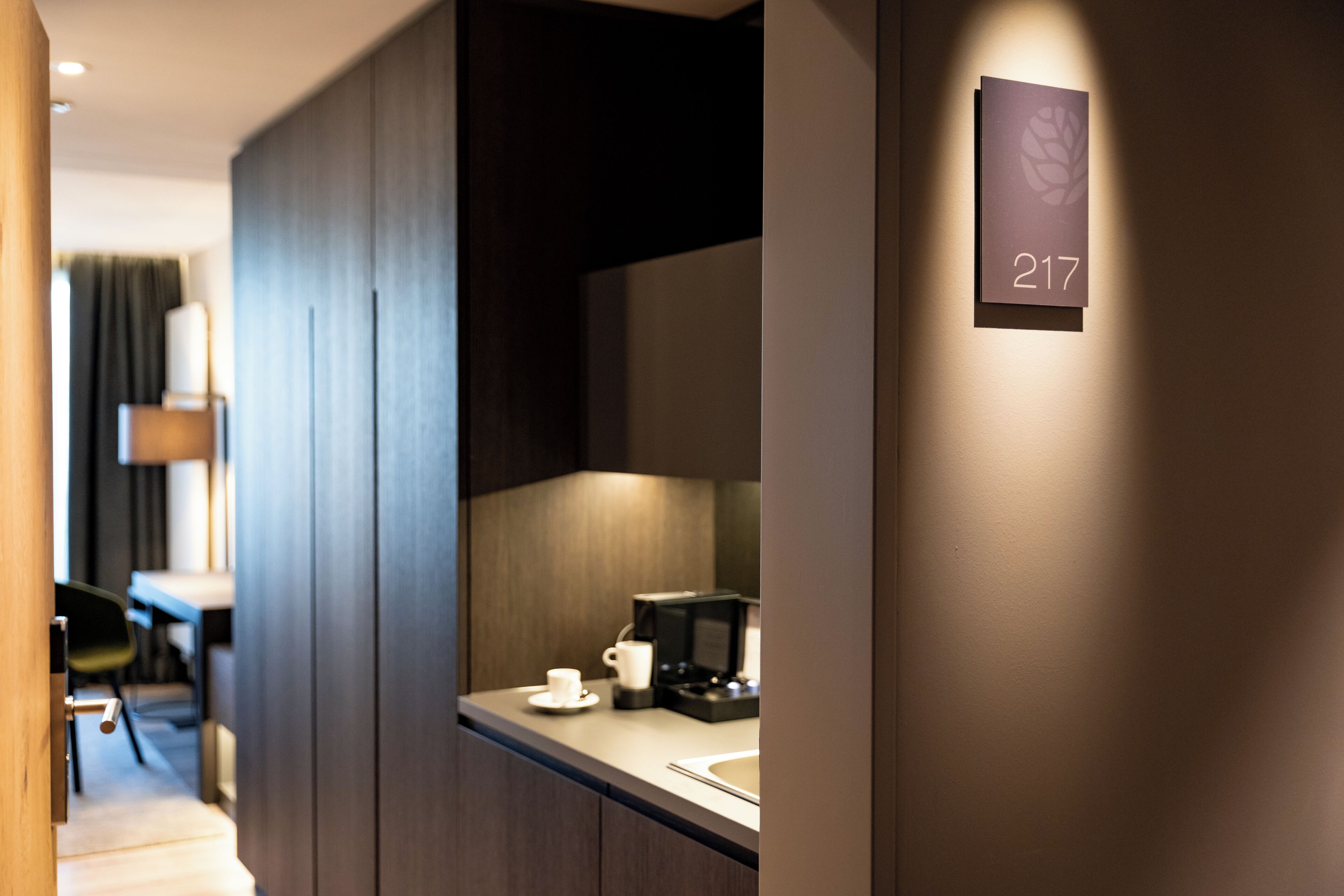 Habitación de hotel con un panel de madera oscura, una pequeña cocina con cafetera y un cartel que indica la habitación 217.
