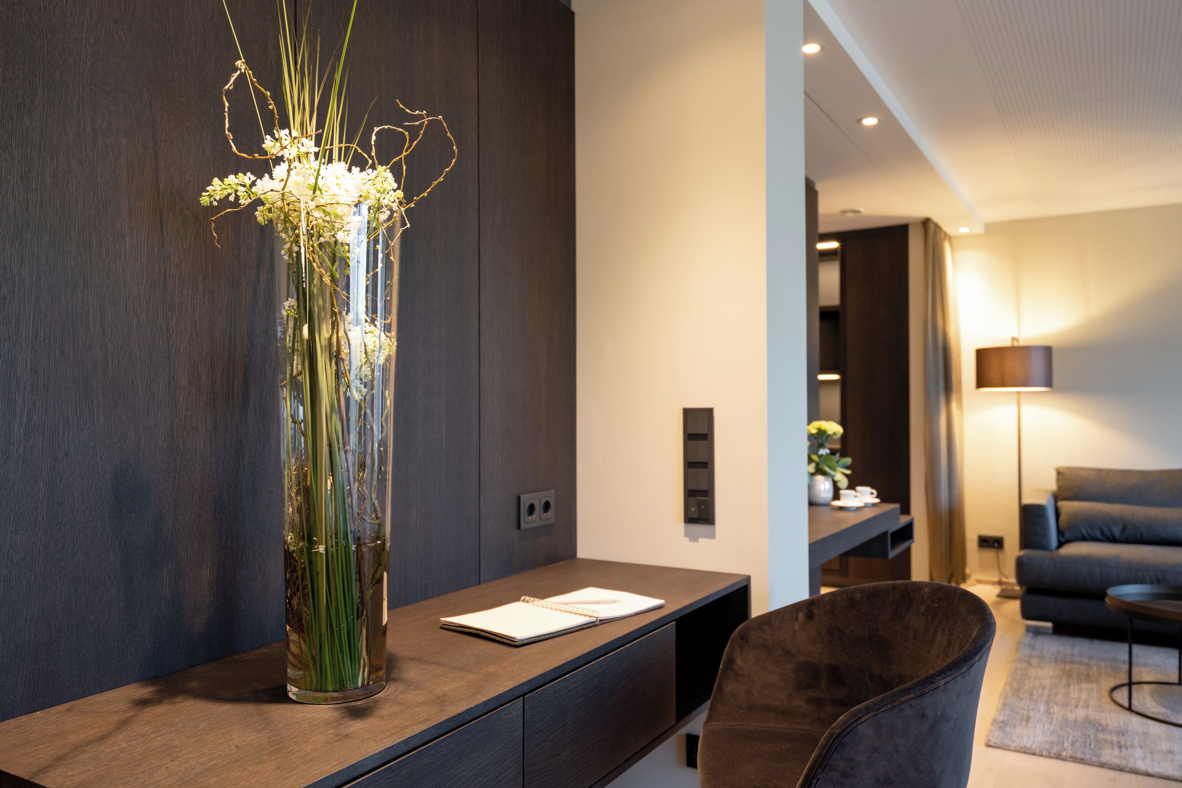 Una sala moderna con una consola de madera oscura, un florero alto con follaje, un sillón de cuero y un sofá oscuro.