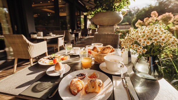 Un espace extérieur aménagé pour prendre les repas, avec des chaises en osier, une table dressée pour le petit-déjeuner, comprenant des croissants, un vase de fleurs et une vue sur le jardin.