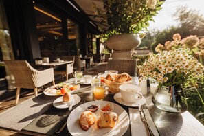 Un espace extérieur aménagé pour prendre les repas, avec des chaises en osier, une table dressée pour le petit-déjeuner, comprenant des croissants, un vase de fleurs et une vue sur le jardin.