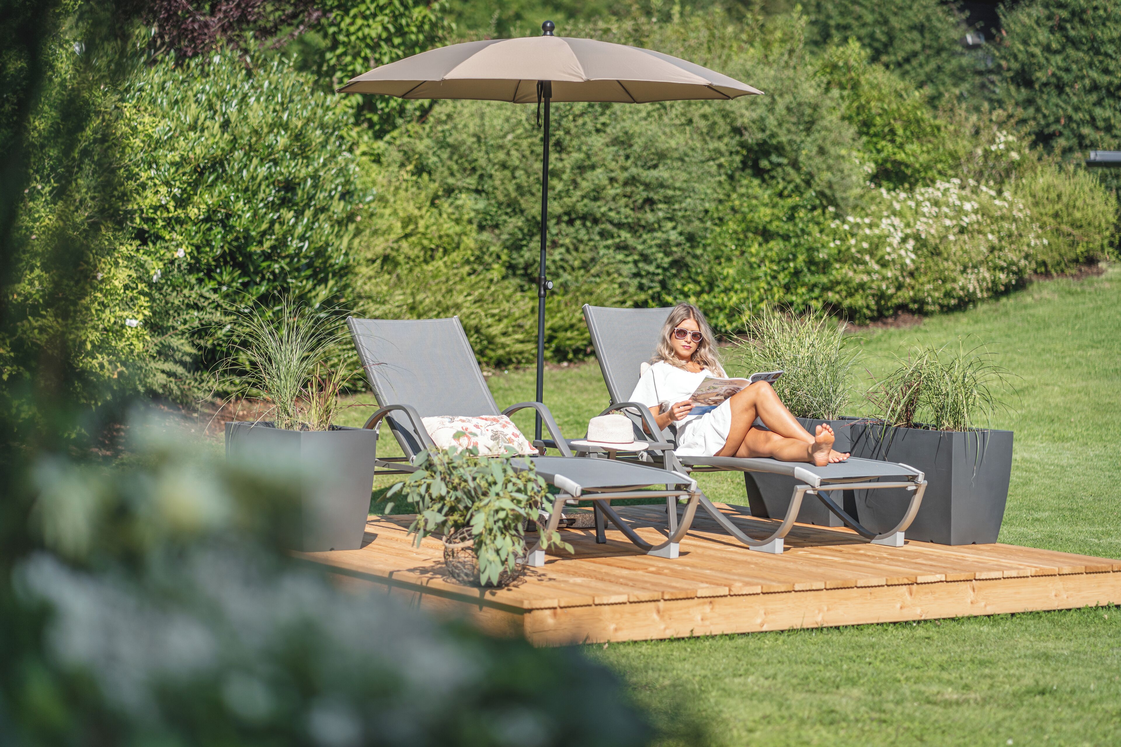 A person relaxing on a sun lounger under a large umbrella, with a garden and greenery in the background.