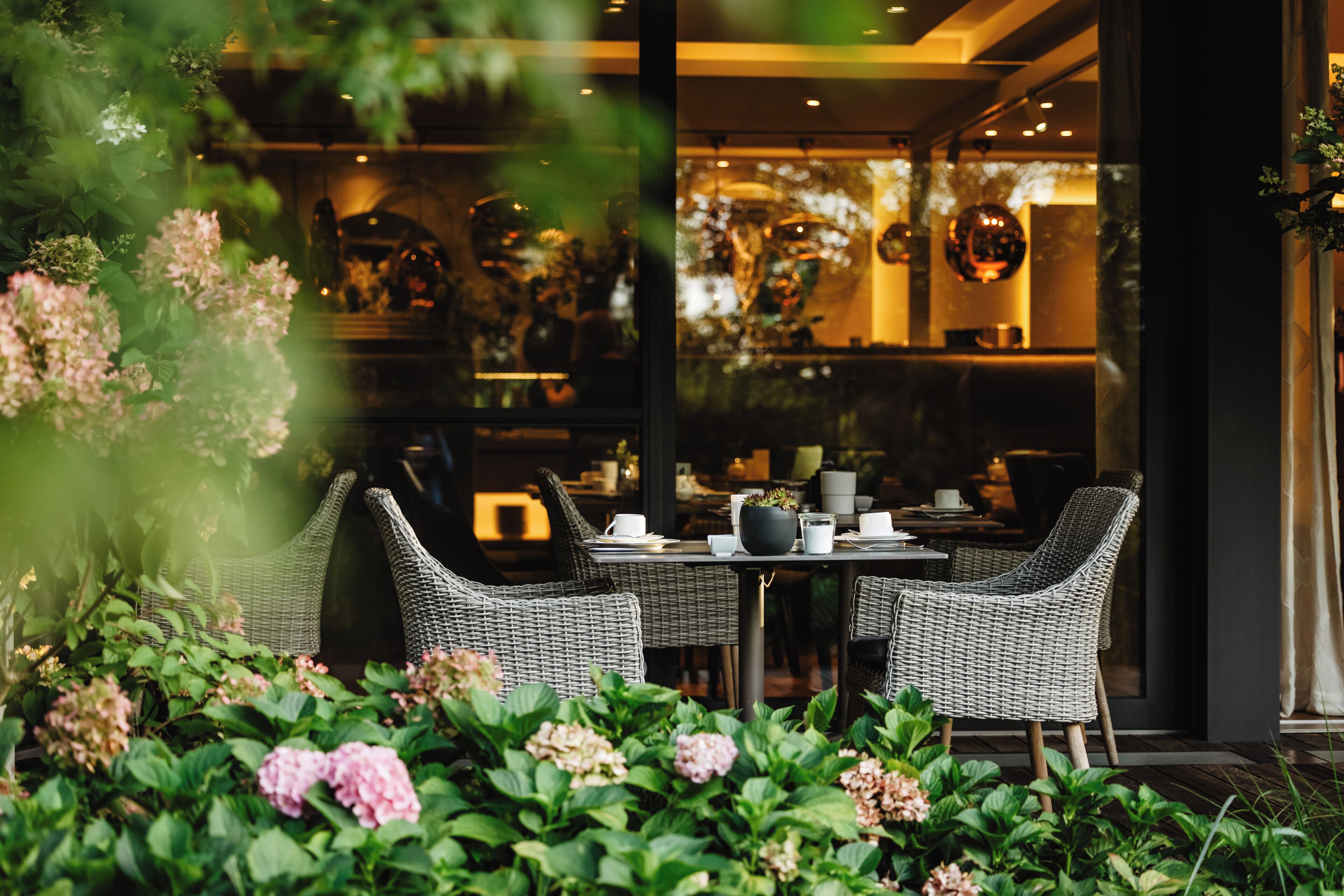 An indoor seating area with wicker chairs and a table, surrounded by greenery and flowers.