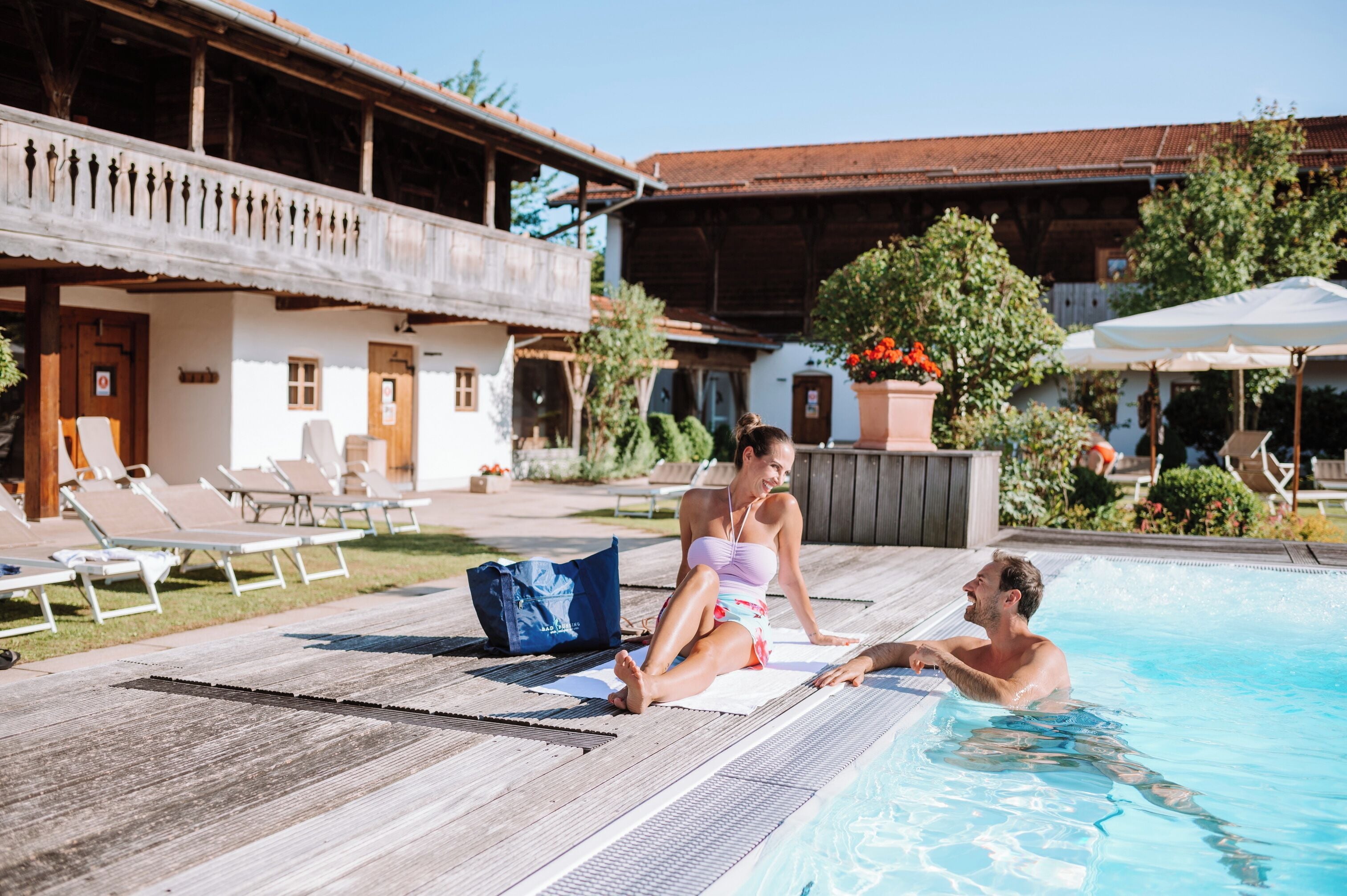 Un área junto a la piscina con sillones de descanso, una terraza de madera y un edificio con balcón.