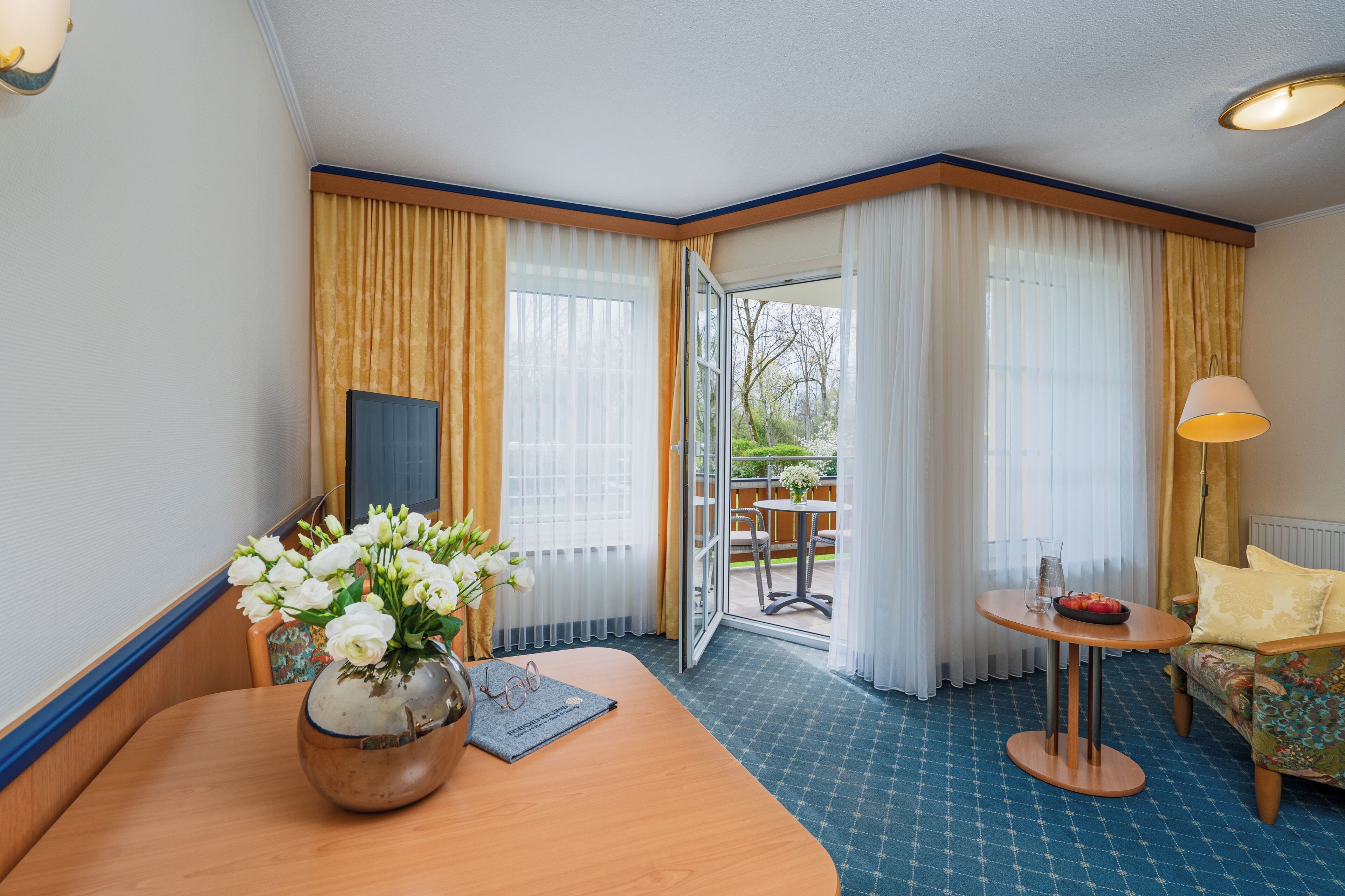 A hotel room with a wooden table, a vase of white flowers, a small round table with a lamp, and a sofa.