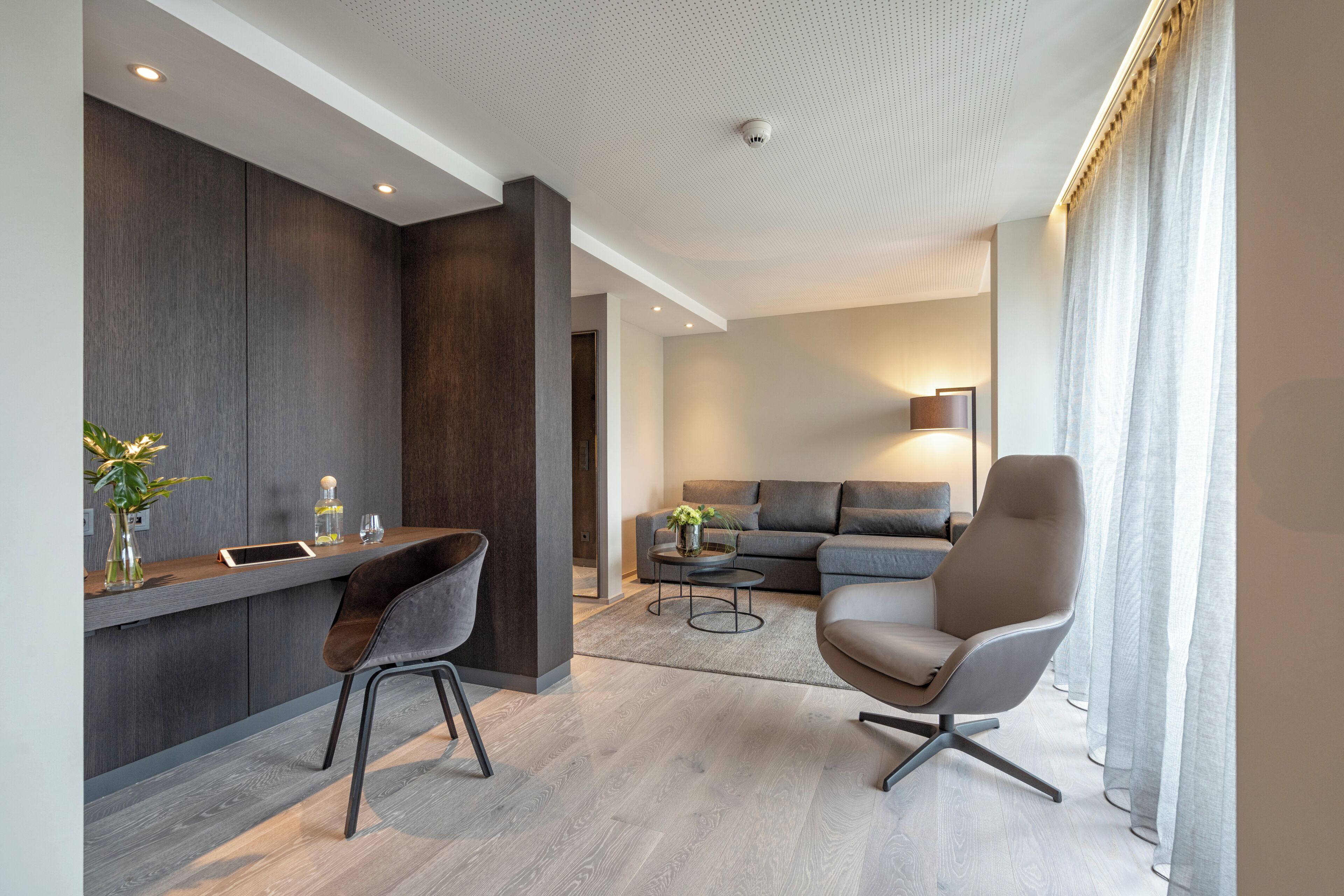 A modern living room with a sofa, armchair, and coffee table. There is a desk with a chair and a vase with a plant.