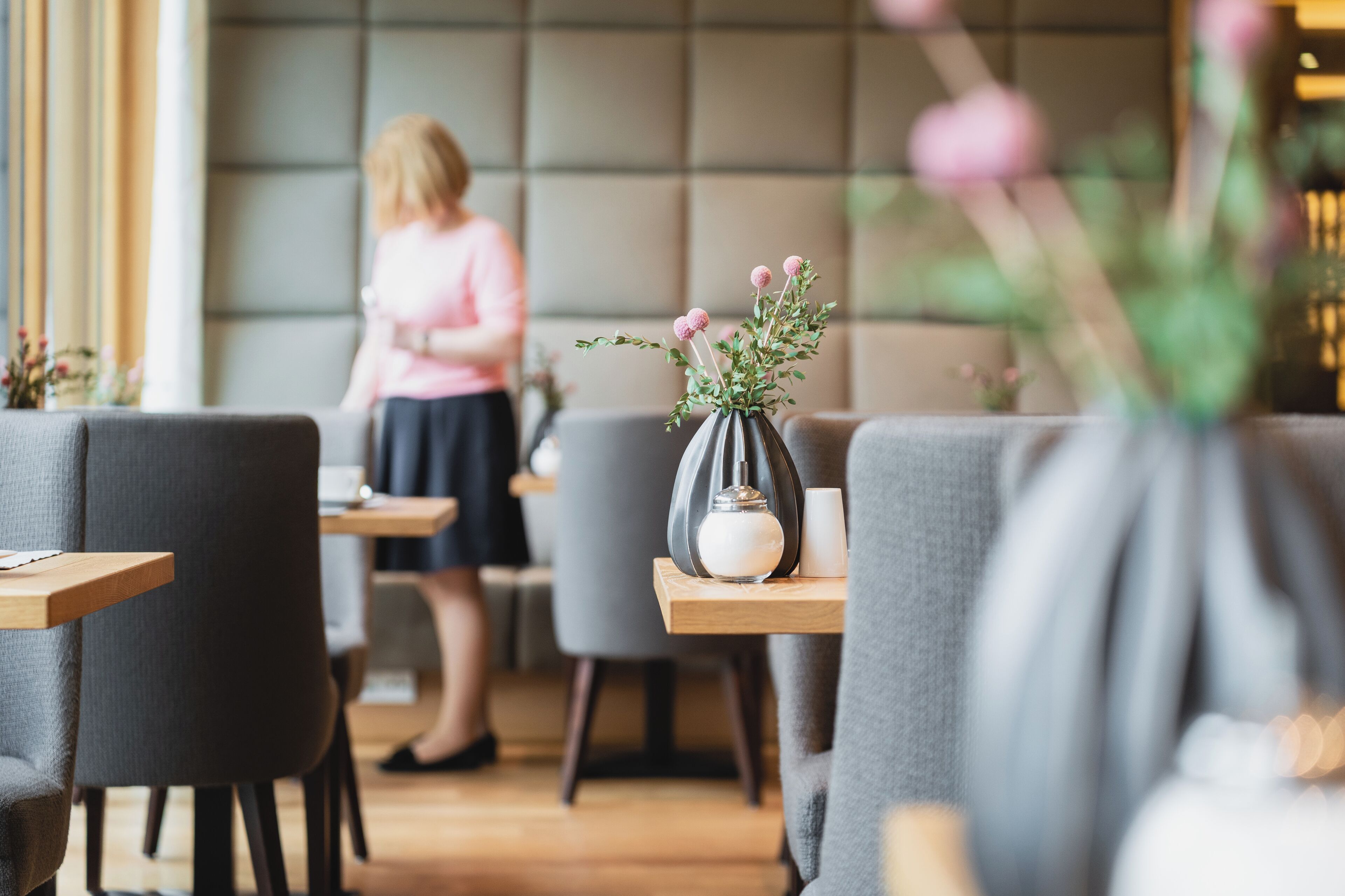Een modern caféinterieur met grijs gestoffeerde stoelen, houten tafels en een vaas met roze bloemen op een tafel.