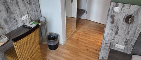 Cocina compacta con suelo de madera, una silla de mimbre, un cesto de basura y una pequeña mesa con un control remoto.
