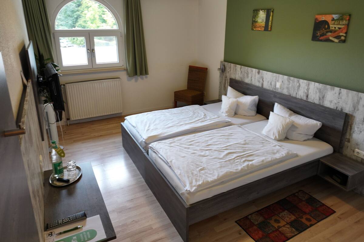 A hotel room with two beds, a window with curtains, a radiator, a chair, and a small table with a bottle and glasses.