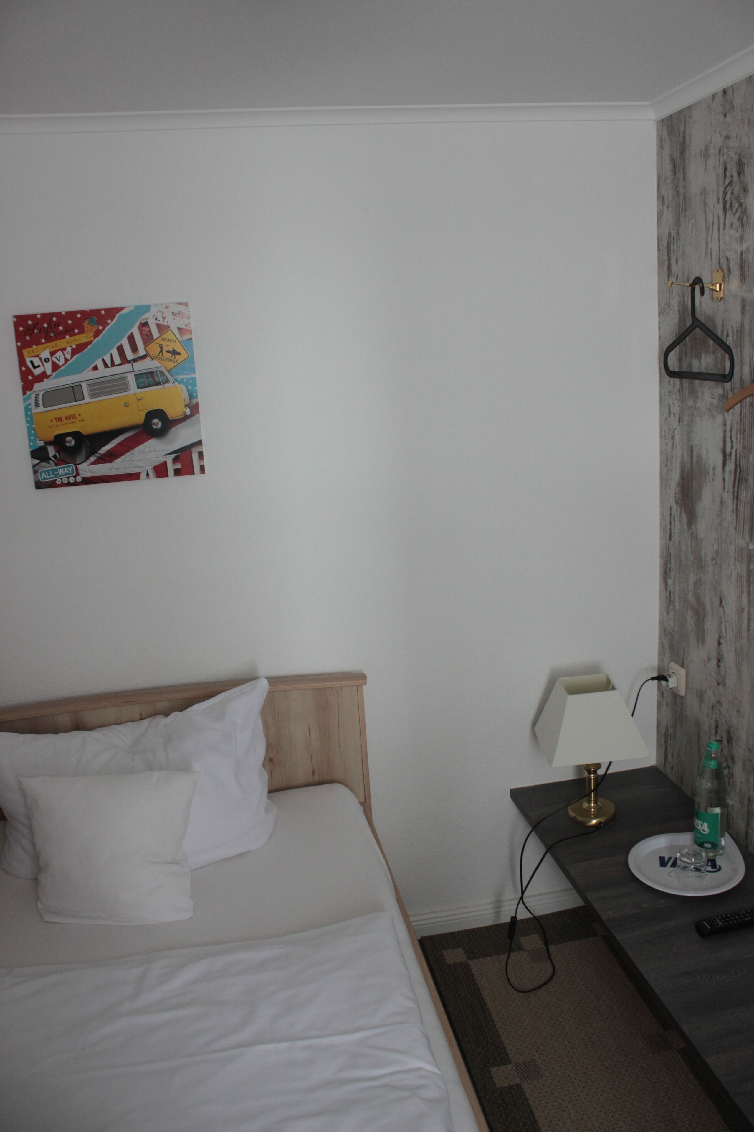 A hotel room with a bed, a nightstand with a lamp, a bottle, and a remote control.