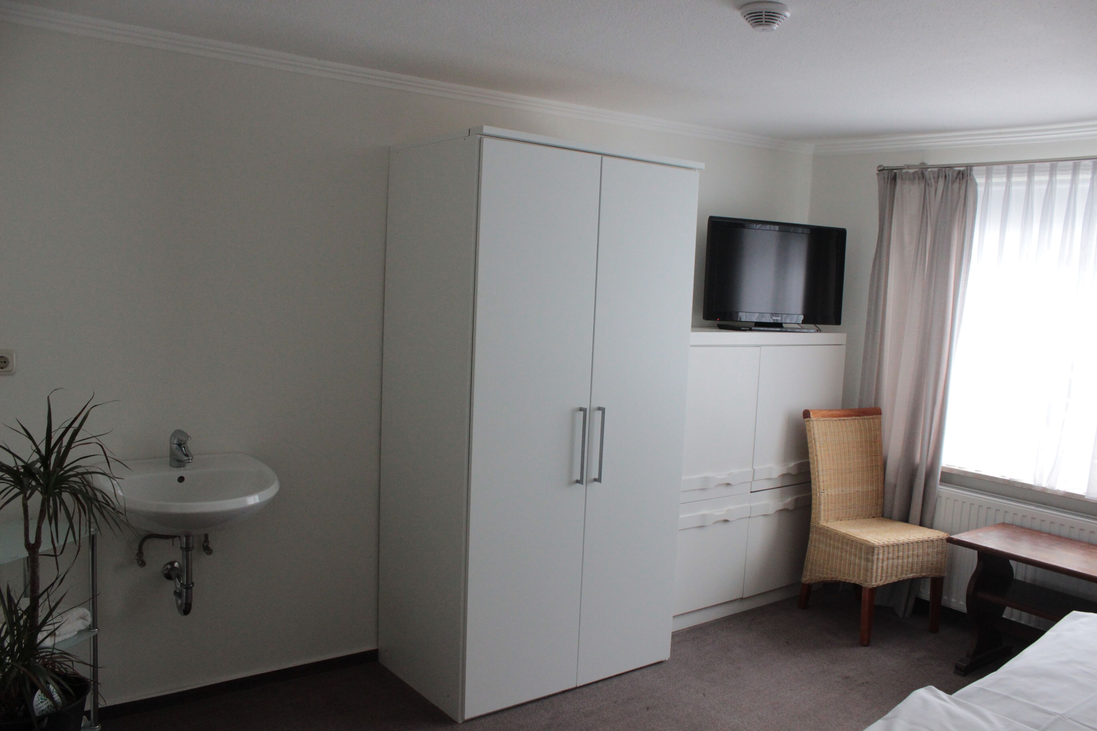 Ein Hotelzimmer mit einem weißen Kleiderschrank, einem an einem Schreibtisch montierten Fernseher, einem Korbstuhl, einem weißen Waschbecken und einer Topfpflanze.