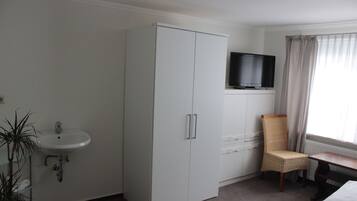 Ein Hotelzimmer mit einem weißen Kleiderschrank, einem an einem Schreibtisch montierten Fernseher, einem Korbstuhl, einem weißen Waschbecken und einer Topfpflanze.
