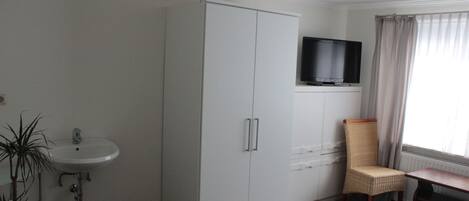 Habitación de hotel con un armario blanco, un televisor sobre un escritorio, una silla de mimbre, un lavabo blanco y una planta en maceta.