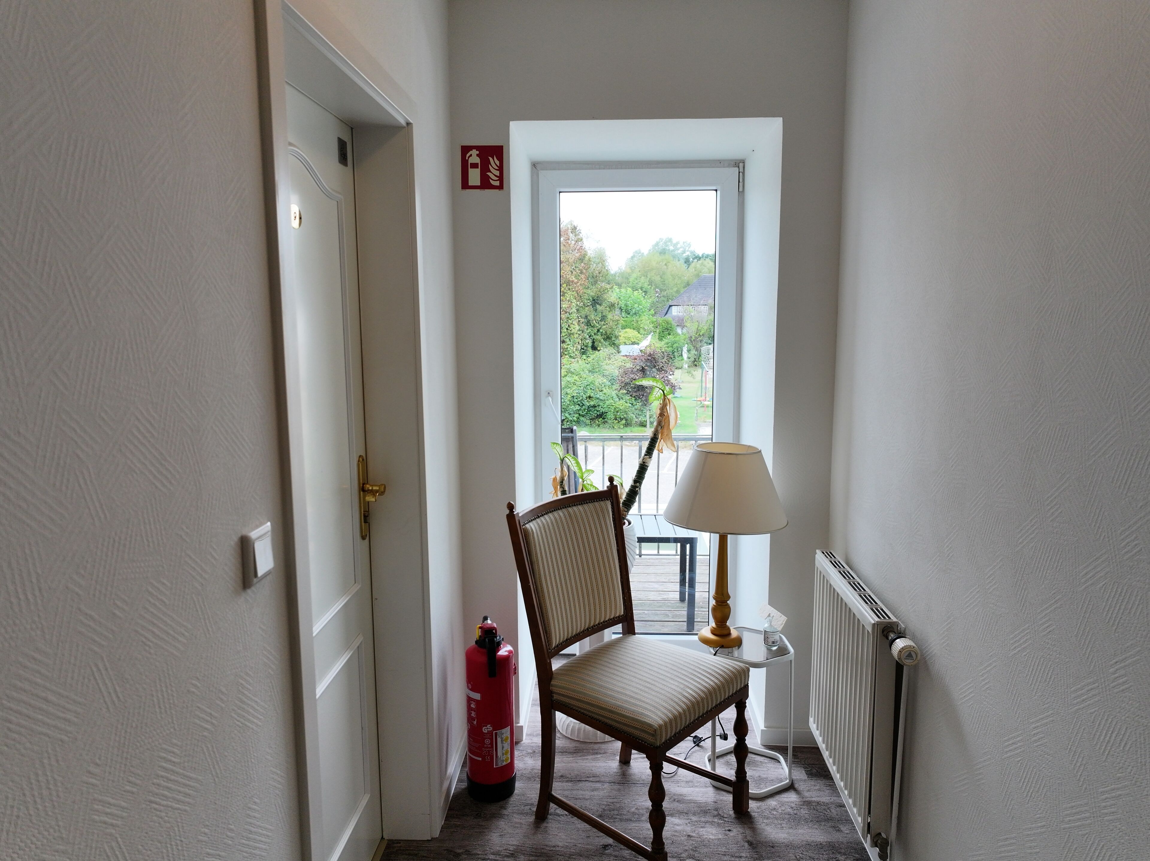 A narrow hallway with a chair and a lamp near a window, a fire extinguisher, and a radiator.