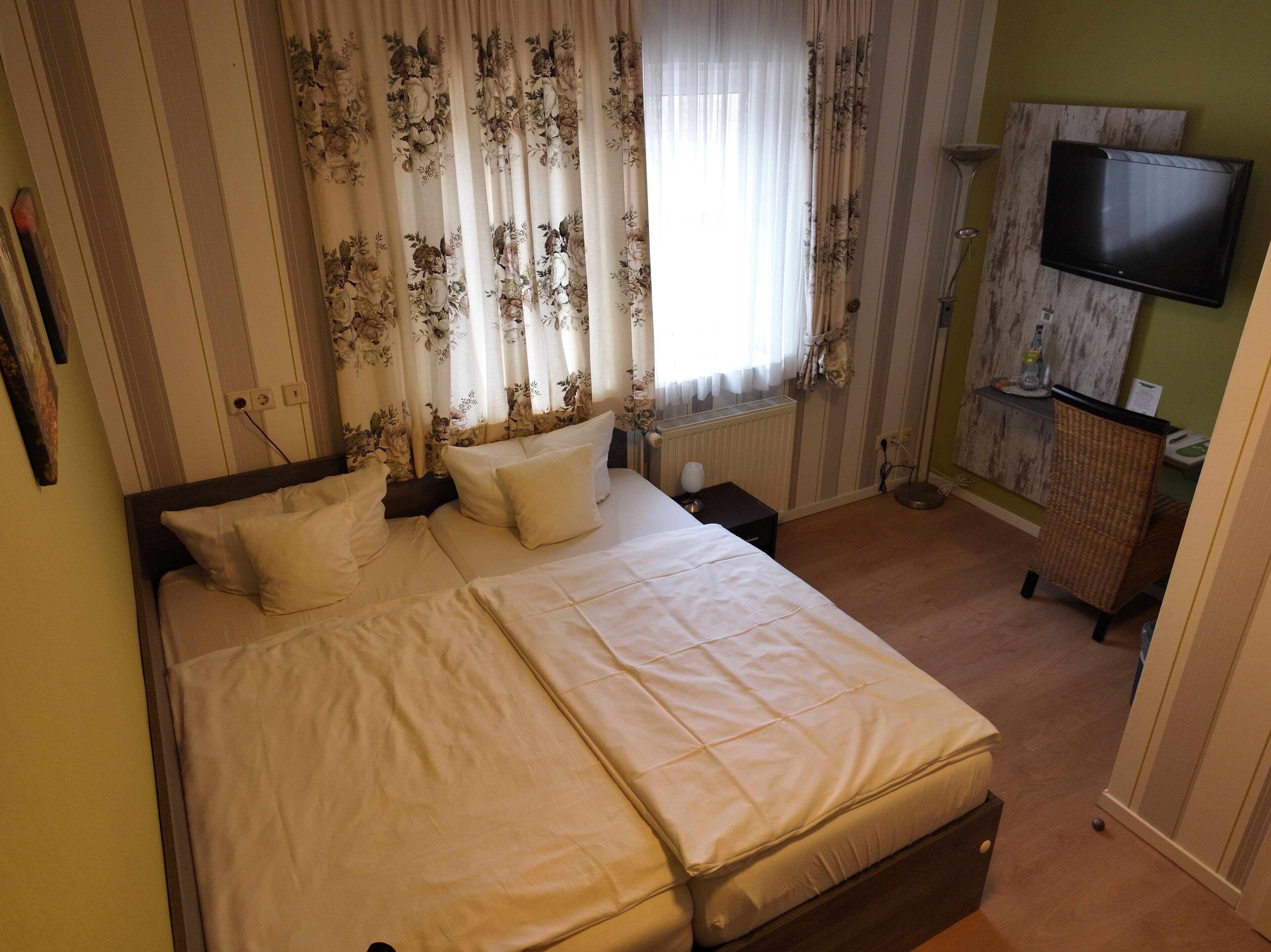 A neatly made bed with white linens and floral curtains, a flat-screen TV mounted on the wall, and a wicker chair.