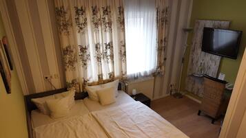 A neatly made bed with white linens and floral curtains, a flat-screen TV mounted on the wall, and a wicker chair.
