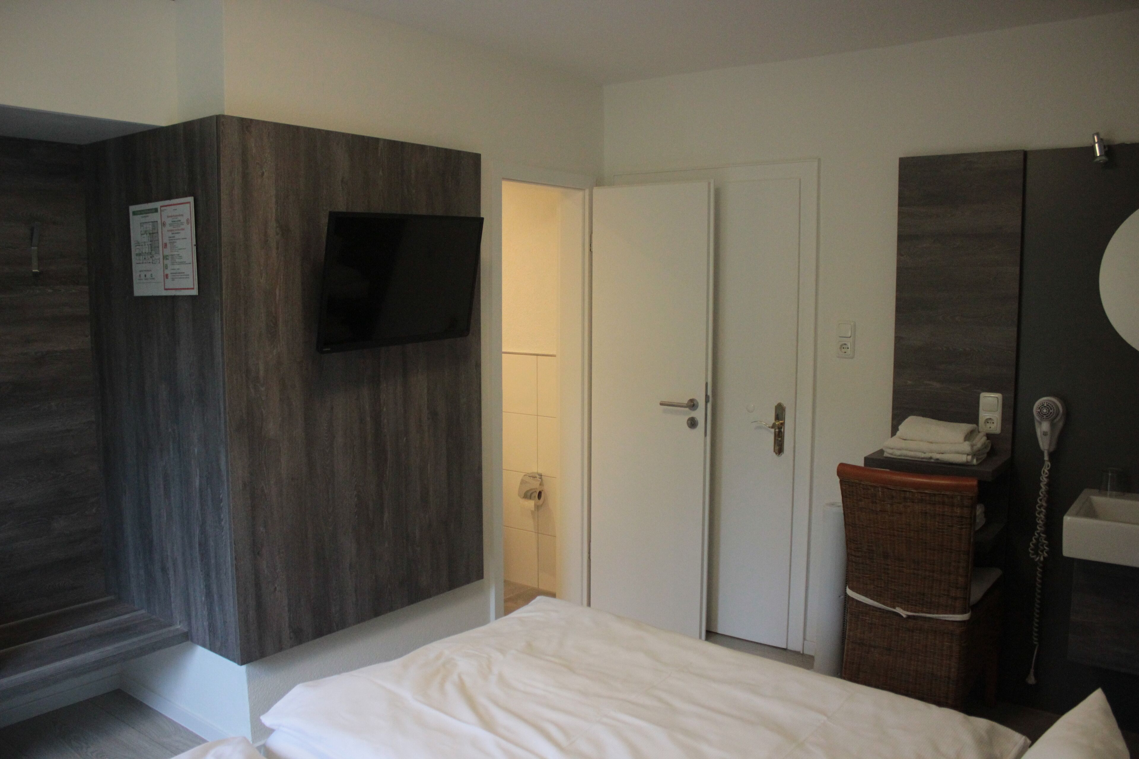 Un dormitorio con cama, televisor montado en la pared, armario, puerta, silla de mimbre y baño con lavamanos y secador de pelo.