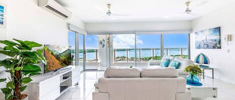A modern living room with a white sofa, a flat-screen TV, and a large window overlooking a beach.
