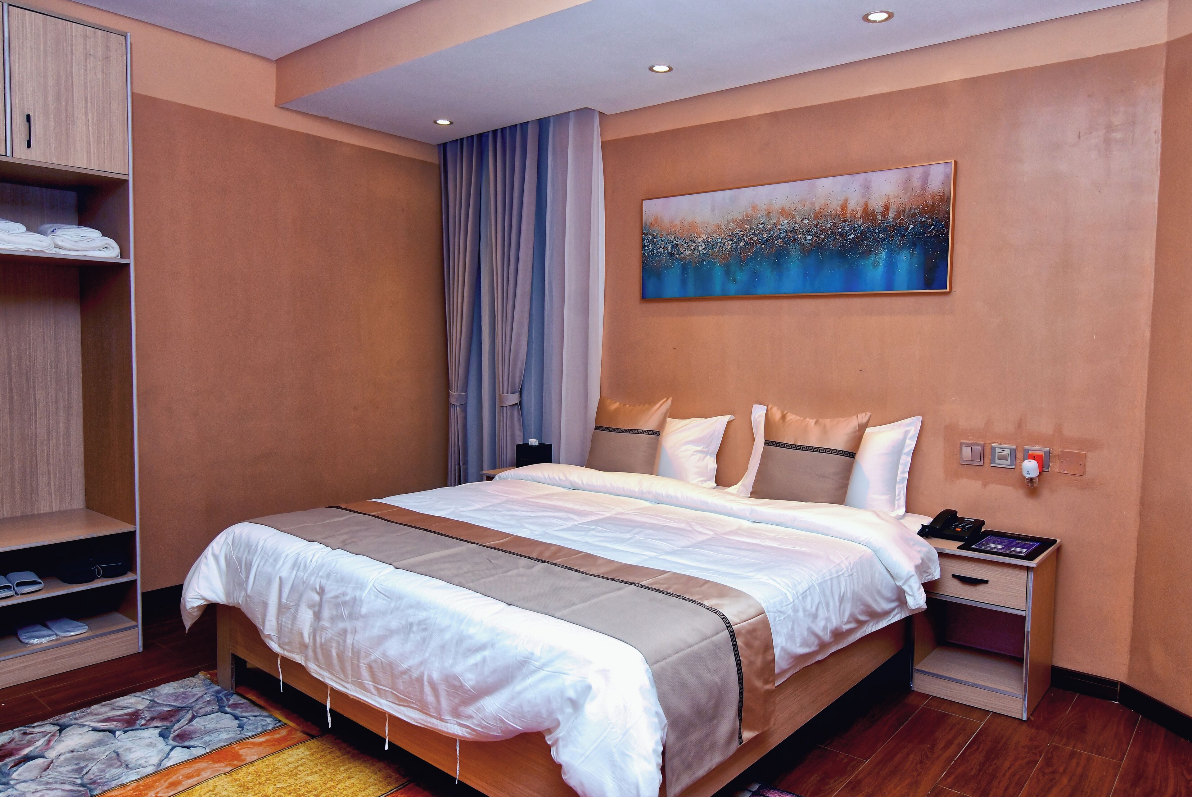 A hotel room with a large bed, a bedside table with a phone, a nightstand, a wardrobe, and a painting on the wall.