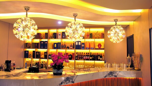 Uma área de bar com bancada de mármore, um vaso de flores cor-de-rosa e prateleiras com garrafas de bebidas.