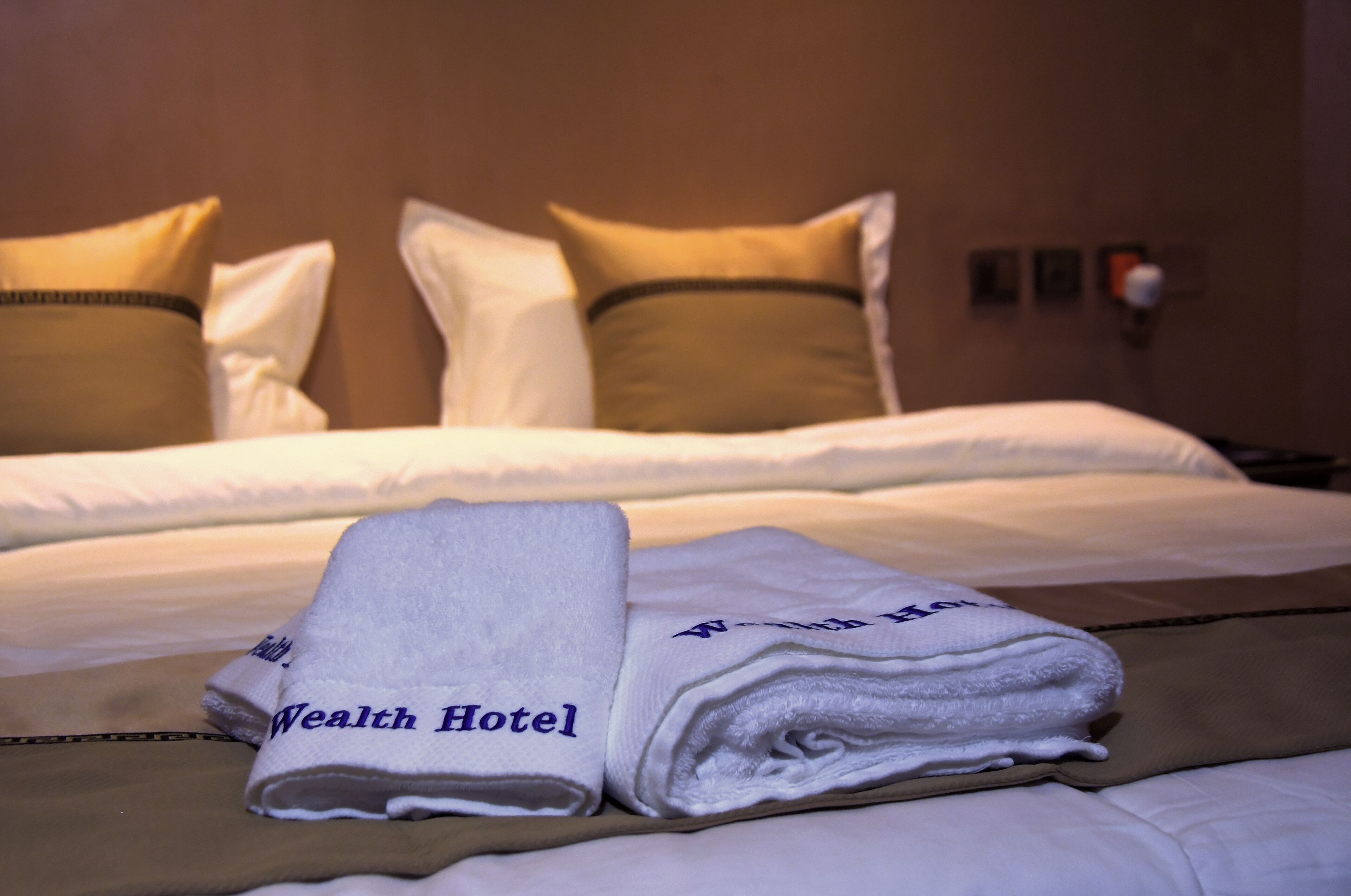 Cama bem arrumada com lençóis brancos e duas almofadas, uma delas com um detalhe dourado. Uma toalha dobrada com a inscrição "Wealth Hotel" bordada está sobre a cama.