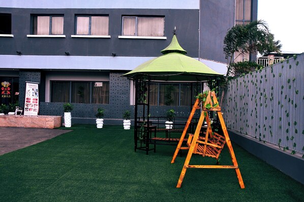 A courtyard with artificial grass, a wooden swing set, a green-roofed gazebo, and a building with large windows and a grey facade. - Wealth Hotel (Lagos)