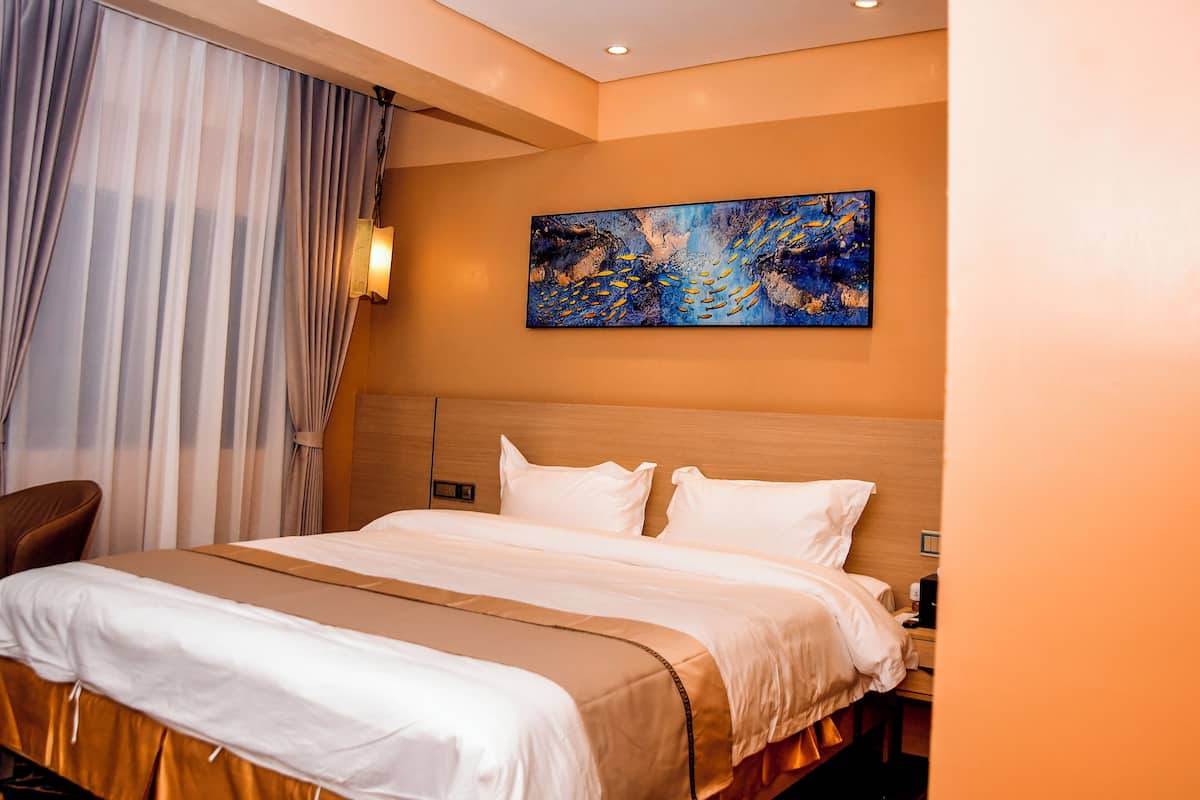 A hotel room with a large bed, a headboard, a painting on the wall, and a window with curtains.