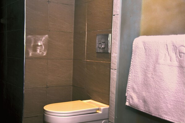 A bathroom with a toilet, a towel hanging on the wall, and a shower area. - Wealth Hotel (Lagos)