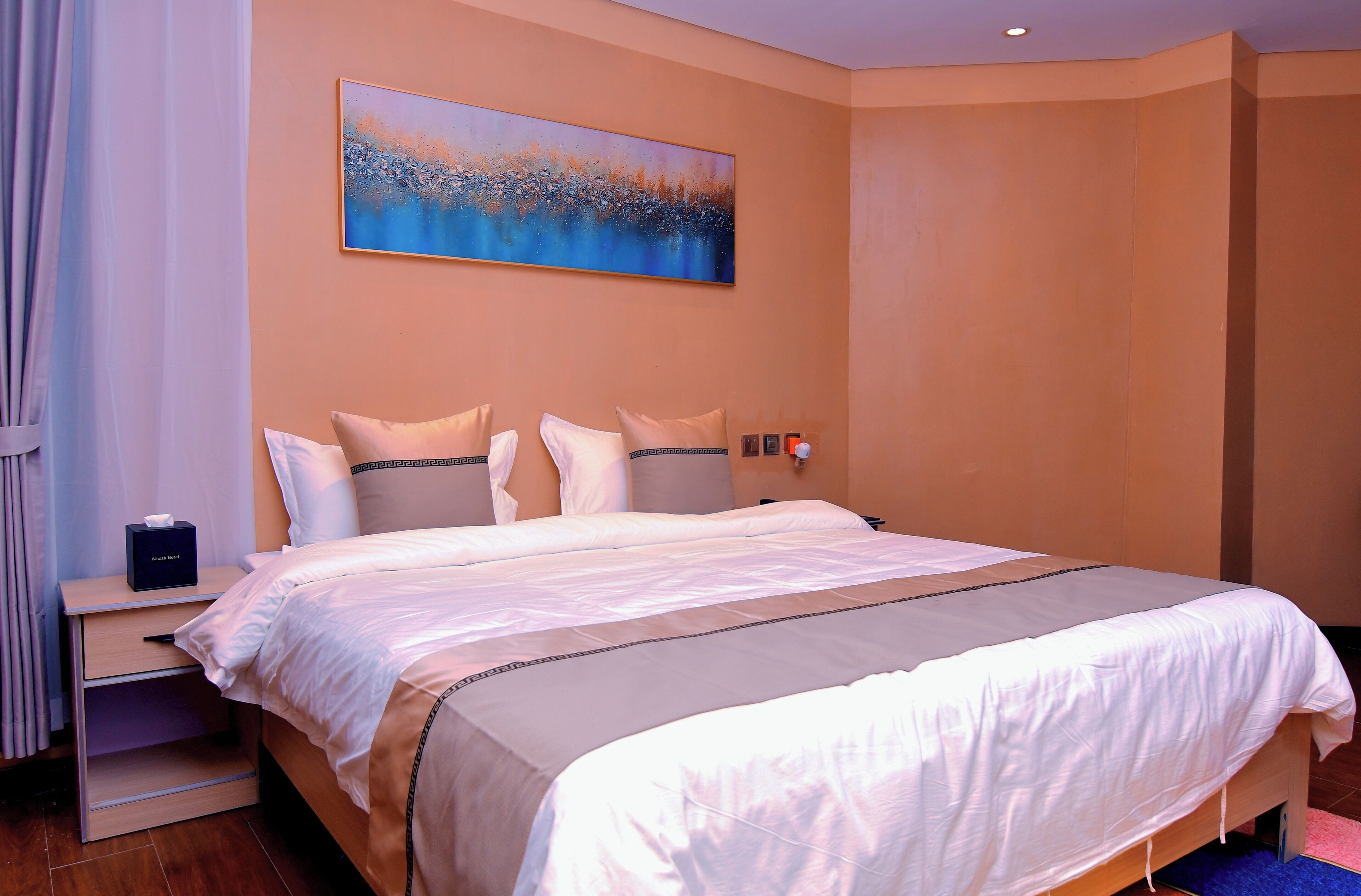 A neatly made bed with a white and beige bedspread, a nightstand with a tissue box, and a painting of a landscape on the wall.