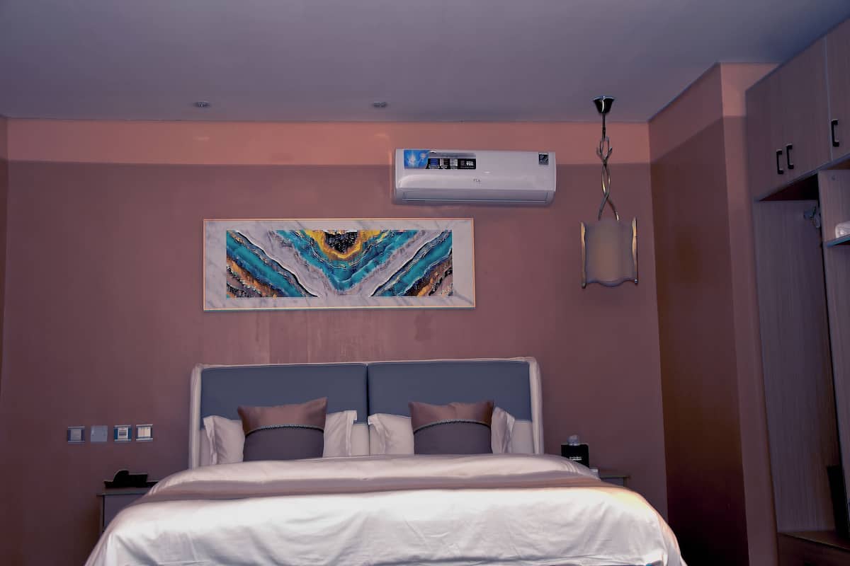 A hotel room with a bed, a painting, an air conditioning unit, and a nightstand.